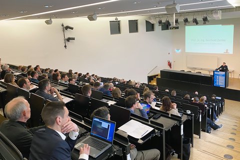 Großer Hörsaal mit ansteigenden Sitzreihen, in denen zahlreiche Teilnehmende sitzen. Vorn steht ein Redner am Pult, hinter ihm ist eine Präsentationsfolie auf eine Leinwand projiziert.