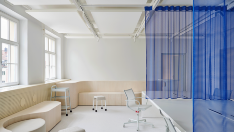 eller Arbeitsbereich mit weißen Schreibtischen, hellen Holzbänken und blauen Vorhängen als Raumteiler vor großen Fenstern.