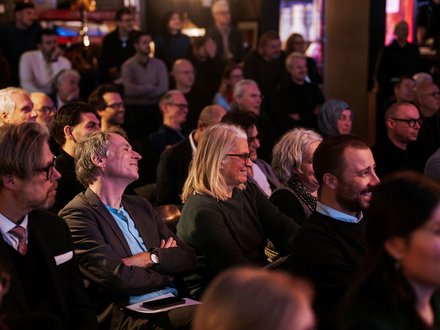Das Bild zeigt das Publikum eines Events aus seitlicher Perspektive. Viele Menschen sitzen dicht nebeneinander in Reihen und schauen aufmerksam nach vorne. Einige lächeln oder wirken interessiert, andere hören konzentriert zu. Die Beleuchtung ist warm und gedämpft, was auf eine Abendveranstaltung oder ein Kultur- bzw. Fachformat hinweist. Zweck des Bildes ist es, die hohe Beteiligung und das Interesse des Publikums zu zeigen und die Atmosphäre einer gut besuchten Veranstaltung einzufangen.