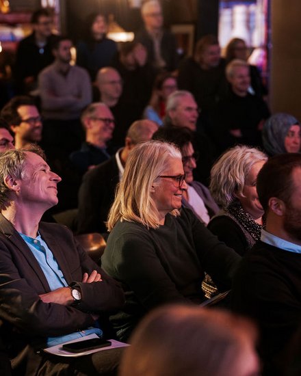 Das Bild zeigt das Publikum eines Events aus seitlicher Perspektive. Viele Menschen sitzen dicht nebeneinander in Reihen und schauen aufmerksam nach vorne. Einige lächeln oder wirken interessiert, andere hören konzentriert zu. Die Beleuchtung ist warm und gedämpft, was auf eine Abendveranstaltung oder ein Kultur- bzw. Fachformat hinweist. Zweck des Bildes ist es, die hohe Beteiligung und das Interesse des Publikums zu zeigen und die Atmosphäre einer gut besuchten Veranstaltung einzufangen.