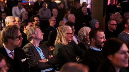 Das Bild zeigt das Publikum eines Events aus seitlicher Perspektive. Viele Menschen sitzen dicht nebeneinander in Reihen und schauen aufmerksam nach vorne. Einige lächeln oder wirken interessiert, andere hören konzentriert zu. Die Beleuchtung ist warm und gedämpft, was auf eine Abendveranstaltung oder ein Kultur- bzw. Fachformat hinweist. Zweck des Bildes ist es, die hohe Beteiligung und das Interesse des Publikums zu zeigen und die Atmosphäre einer gut besuchten Veranstaltung einzufangen.
