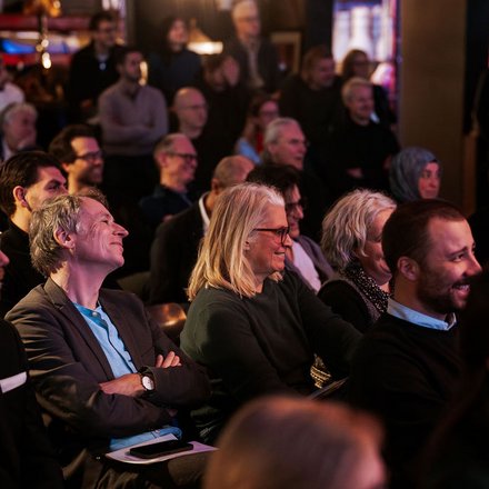 Das Bild zeigt das Publikum eines Events aus seitlicher Perspektive. Viele Menschen sitzen dicht nebeneinander in Reihen und schauen aufmerksam nach vorne. Einige lächeln oder wirken interessiert, andere hören konzentriert zu. Die Beleuchtung ist warm und gedämpft, was auf eine Abendveranstaltung oder ein Kultur- bzw. Fachformat hinweist. Zweck des Bildes ist es, die hohe Beteiligung und das Interesse des Publikums zu zeigen und die Atmosphäre einer gut besuchten Veranstaltung einzufangen.
