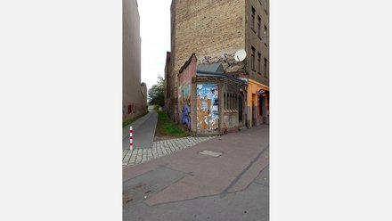 Kleines Eckgebäude vor der Renovierung mit Graffiti, Plakaten und verwitterter Fassade an einer schmalen Straße neben einem Radweg.