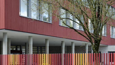 Detailansicht einer roten Gebäudefassade hinter einem farbenfrohen Lamellenzaun in Rot-, Orange- und Gelbtönen mit einem Baum im Vordergrund.
