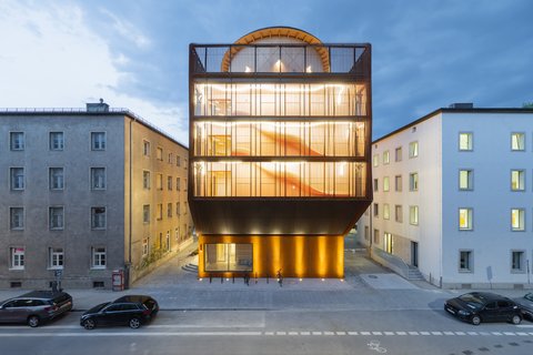 Modernes, mehrstöckiges Gebäude mit großflächigen Glasfronten und beleuchteter Fassade zwischen zwei älteren Gebäuden an einer Straße mit parkenden Autos