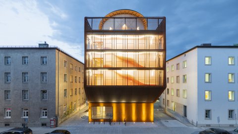 Modernes, mehrstöckiges Gebäude mit großflächigen Glasfronten und beleuchteter Fassade zwischen zwei älteren Gebäuden an einer Straße mit parkenden Autos