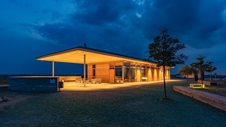 Flaches Besucherzentrum aus Holz/Stampflehm, abends warm beleuchtet unter weitem Vordach vor dunklem Himmel.