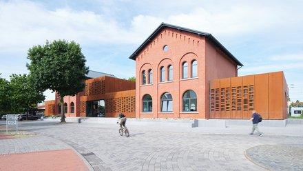 Modernisiertes Bahnhofsgebäude  Ein ehemaliges Backsteingebäude eines Bahnhofs wurde modernisiert und durch seitliche, rostfarbene Metallfassaden ergänzt. Große Fenster und ein zentraler Eingang öffnen das Gebäude zum Vorplatz. Auf dem Platz davor bewegen sich Menschen zu Fuß und mit dem Fahrrad. Das Bild zeigt die Verbindung von historischer Bausubstanz und zeitgenössischer Architektur. Es steht für die Umnutzung und Aufwertung von Bahnhöfen als öffentliche Orte.