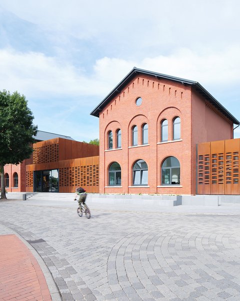 Modernisiertes Bahnhofsgebäude  Ein ehemaliges Backsteingebäude eines Bahnhofs wurde modernisiert und durch seitliche, rostfarbene Metallfassaden ergänzt. Große Fenster und ein zentraler Eingang öffnen das Gebäude zum Vorplatz. Auf dem Platz davor bewegen sich Menschen zu Fuß und mit dem Fahrrad. Das Bild zeigt die Verbindung von historischer Bausubstanz und zeitgenössischer Architektur. Es steht für die Umnutzung und Aufwertung von Bahnhöfen als öffentliche Orte.