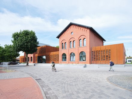 Modernisiertes Bahnhofsgebäude  Ein ehemaliges Backsteingebäude eines Bahnhofs wurde modernisiert und durch seitliche, rostfarbene Metallfassaden ergänzt. Große Fenster und ein zentraler Eingang öffnen das Gebäude zum Vorplatz. Auf dem Platz davor bewegen sich Menschen zu Fuß und mit dem Fahrrad. Das Bild zeigt die Verbindung von historischer Bausubstanz und zeitgenössischer Architektur. Es steht für die Umnutzung und Aufwertung von Bahnhöfen als öffentliche Orte.