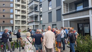 Gruppe von Menschen steht vor modernen Wohngebäuden mit Balkonen und großen Fenstern auf einem Gehweg.