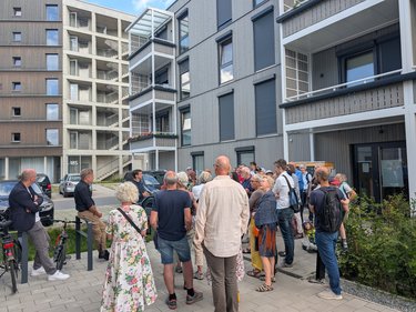Gruppe von Menschen steht vor modernen Wohngebäuden mit Balkonen und großen Fenstern auf einem Gehweg.
