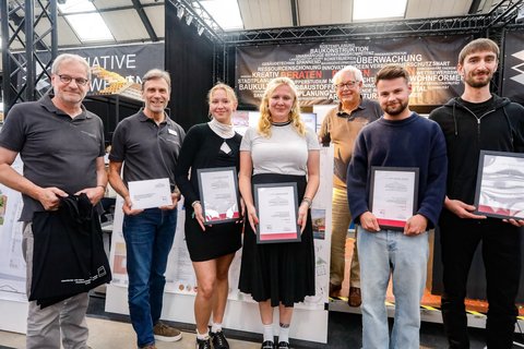 Gruppe von Preisträger:innen mit Urkunden auf einem Messestand der Initiative Bauwesen.