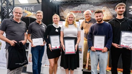 Gruppe von Preisträger:innen mit Urkunden auf einem Messestand der Initiative Bauwesen.