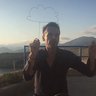 „Ein Mann hockt gestikulierend im Gegenlicht vor einer Berglandschaft, während direkt hinter ihm eine stilisierte Wolke aus Draht in den Abendhimmel ragt.“