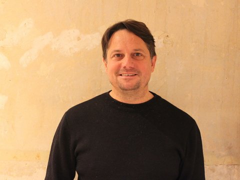 Mann mittleren Alters mit dunklen Haaren lächelt in die Kamera, trägt schwarzes Langarmshirt vor beiger Wand
