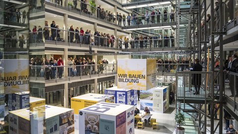 Mehrstöckiges Gebäude mit vielen Menschen auf den Balkonen und Ausstellungsständen im Innenhof, auf einem Banner steht 'WIR BAUEN BILDUNG'