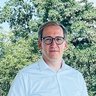 "Mann mit Brille steht auf einer modernen Betonterrasse mit Glasgeländer. Er trägt ein hellblaues Hemd, dunkelblaue Jeans und braune Schuhe. Im Hintergrund sind grüne Bäume, eine Kirchturmspitze und eine Stadt zu sehen. Ein grauer Betonpfeiler und ein Betonvordach rahmen die Szene ein."