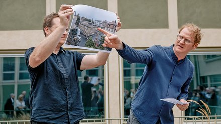 Zwei Männer in blauen Hemden halten gemeinsam ein Foto einer Stadtstraße mit Häusern hoch. Sie stehen vor einem modernen Gebäude mit grünen Fensterfronten. Ein großes Publikum ist im Hintergrund verschwommen sichtbar.