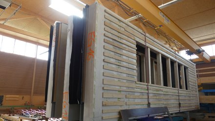 Fabrikhalle: Vorfertigte Holztafelwand mit Fensteröffnungen steht auf einem Wagen; Kranriemen zum Versetzen sind befestigt.