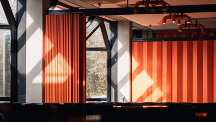 Innenraum mit orangefarbenen Faltwänden und Deckenstrahlern; Sonnenlicht und Spiegelung auf glänzender Oberfläche im Vordergrund.