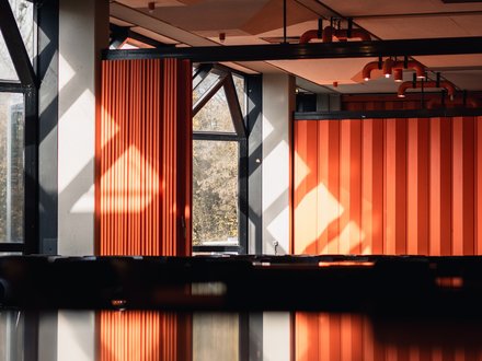 Innenraum mit orangefarbenen Faltwänden und Deckenstrahlern; Sonnenlicht und Spiegelung auf glänzender Oberfläche im Vordergrund.