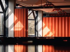 Innenraum mit orangefarbenen Faltwänden und Deckenstrahlern; Sonnenlicht und Spiegelung auf glänzender Oberfläche im Vordergrund.