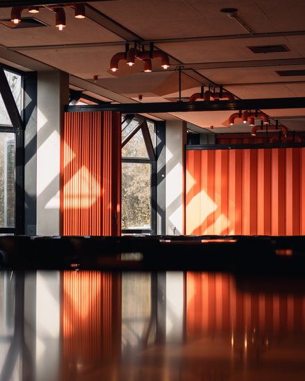 Innenraum mit orangefarbenen Faltwänden und Deckenstrahlern; Sonnenlicht und Spiegelung auf glänzender Oberfläche im Vordergrund.