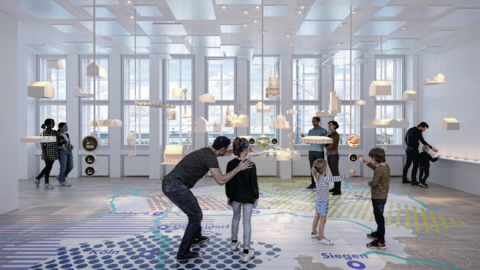 Mehrere Personen, darunter Kinder und Erwachsene, betrachten in einem hellen Raum mit großen Fenstern eine Ausstellung mit schwebenden Modellen und einer großflächigen Bodenkarte mit Städtenamen