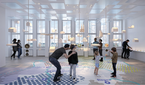 Mehrere Personen, darunter Kinder und Erwachsene, betrachten in einem hellen Raum mit großen Fenstern eine Ausstellung mit schwebenden Modellen und einer großflächigen Bodenkarte mit Städtenamen