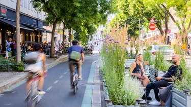 Verkehrsberuhigte Straße mit Fahrradfahrern und Begrünung sowie Sitzgelegenheiten.