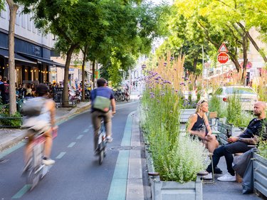 Verkehrsberuhigte Straße mit Fahrradfahrern und Begrünung sowie Sitzgelegenheiten.