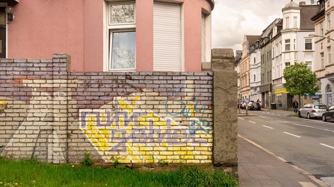 Backsteinmauer mit Graffiti vor einem rosafarbenen Eckhaus an einer städtischen Straßenecke.