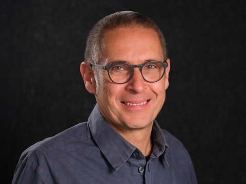 Porträt eines lächelnden Mannes mit Brille vor schwarzem Hintergrund.