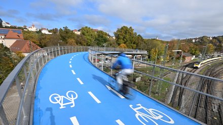 Blick entlang der blauen Radbrücke, ein Radfahrer fährt über die Kurve, Bahnstrecke daneben.