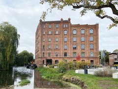 Backsteingebäude an einem Wasserlauf, umgeben von Grünflächen und Bäumen.