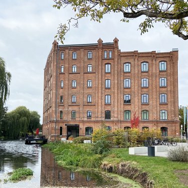 Backsteingebäude an einem Wasserlauf, umgeben von Grünflächen und Bäumen.