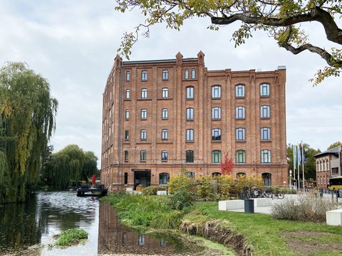 Backsteingebäude an einem Wasserlauf, umgeben von Grünflächen und Bäumen.