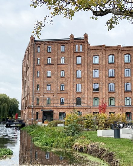 Backsteingebäude an einem Wasserlauf, umgeben von Grünflächen und Bäumen.