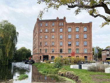 Backsteingebäude an einem Wasserlauf, umgeben von Grünflächen und Bäumen.