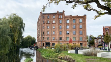 Backsteingebäude an einem Wasserlauf, umgeben von Grünflächen und Bäumen.