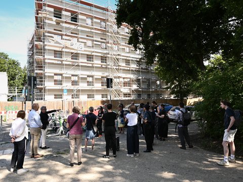 "Etwa 20 Personen stehen im Halbkreis auf sandigem Untergrund vor einem mehrstöckigen Neubau. Das weiße Wohngebäude mit Holz- und Metallfassadenelementen ist mit Bauzäunen und orangenen Sicherheitsmarkierungen umgeben. Links grüne Vegetation und Bäume. Im Vordergrund ein Fahrrad. Sonnige Tageslichtsituation. Banner mit Text 'Architektur' sichtbar."