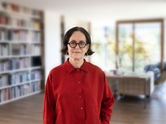"Porträt einer Frau mittleren Alters mit schulterlangenm dunklem Haar und Brille mit schwarzem Rahmen. Sie trägt einen leuchtend roten Anzug aus Hemd und Hose mit Perlmuttknöpfen. Sie steht aufrecht in einem hellen Wohnraum mit Holzboden. Im Hintergrund sind ein beige gefülltes Bücherregal auf der linken Seite und großflächige Fenster mit braunem Holzrahmen auf der rechten Seite sichtbar. Ein Beistelltisch und weitere Einrichtungselemente sind unscharf im Hintergrund erkennbar."