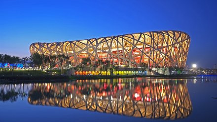 Das beleuchtete Pekinger Nationalstadion "Vogelnest" bei Nacht. Das oval geformte Gebäude mit charakteristischem Stahlgitterwerk ist in Gelb-, Orange- und Rottönen beleuchtet. Das Stadion spiegelt sich in einem ruhigen Gewässer davor. Ein klarer blauer Himmel ist zu sehen.