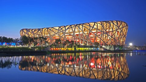 Das beleuchtete Pekinger Nationalstadion "Vogelnest" bei Nacht. Das oval geformte Gebäude mit charakteristischem Stahlgitterwerk ist in Gelb-, Orange- und Rottönen beleuchtet. Das Stadion spiegelt sich in einem ruhigen Gewässer davor. Ein klarer blauer Himmel ist zu sehen.