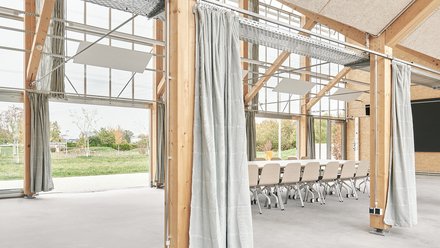 Innenraumdetail mit Holzstützen und Vorhängen, glattem Boden und Blick ins Grüne; Besprechungstisch im Hintergrund.