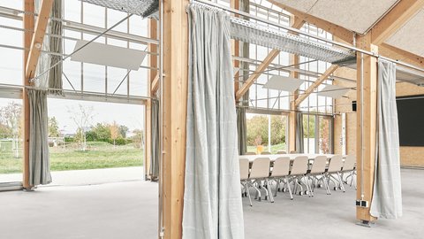 Innenraumdetail mit Holzstützen und Vorhängen, glattem Boden und Blick ins Grüne; Besprechungstisch im Hintergrund.