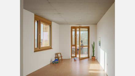 Heller Wohnraum mit Holzfenstern und geöffneter Balkontür, Terrakottafliesen; eine Person sitzt draußen. Lang Schlichter Innenraum mit weißen Wänden und rötlich-braunen Fliesen. Links großes Holzfenster, vorn geöffnete Balkontür zum Balkon. Wenige Gegenstände (Hocker, Rollschuhe, Ball, Zimmerpflanze). Auf dem Balkon sitzt eine Person.