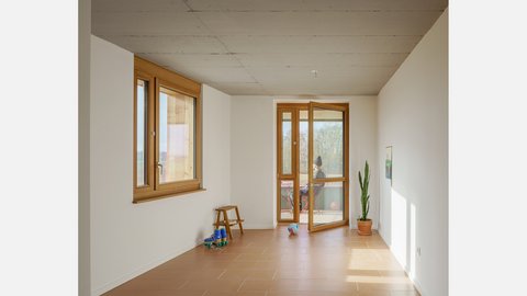 Heller Wohnraum mit Holzfenstern und geöffneter Balkontür, Terrakottafliesen; eine Person sitzt draußen. Lang Schlichter Innenraum mit weißen Wänden und rötlich-braunen Fliesen. Links großes Holzfenster, vorn geöffnete Balkontür zum Balkon. Wenige Gegenstände (Hocker, Rollschuhe, Ball, Zimmerpflanze). Auf dem Balkon sitzt eine Person.