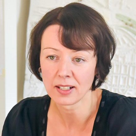 Frau mit brünetten Haaren und Pony sitzt an einem Holztisch in einem Büro. Sie trägt ein schwarzes Kurzarmshirt und gestikuliert mit beiden Händen während sie spricht. Im Hintergrund sind ein Whiteboard und eine Stadtplan-Karte an der Wand sichtbar.